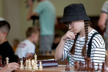A young woman in glasses concentrates on a move while playing chess. A girl plays in a chess tournament.  Girl at the chessboard. Tournament, portrait of teenage girl playing chess	
