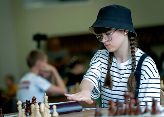 A young woman in glasses concentrates on a move while playing chess. A girl plays in a chess tournament.  Girl at the chessboard. Tournament, portrait of teenage girl playing chess	