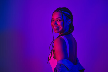 Lovely woman with braided hair in neon lighting, wearing a denim jacket and smiling confidently...