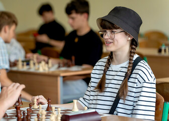 A young woman in glasses concentrates on a move while playing chess. A girl plays in a chess tournament.  Girl at the chessboard. Tournament, portrait of teenage girl playing chess	