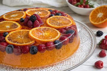 shows a fruit gelatin dessert topped with slices of orange, raspberries, and blueberries, placed on a glass plate on a light wooden table. There is a bowl of fresh berries in the background and a half