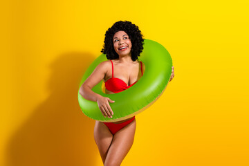 Young woman posing in swimwear with inflatable ring on vivid yellow background, expressing joy and...