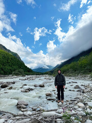 Tourist joyful man stands on rocky bank of mountain river against mountain landscape with glacier and green slopes. Nature of North Ossetia, nature reserve