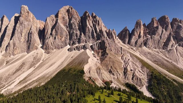 Breathtaking drone shot of dramatic Dolomite peaks rising above lush alpine forests and green valleys. Sunlight grazes the rocky summits of northern Italy's iconic UNESCO mountains