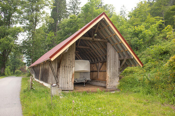 Wooden shed with a red metal roof storing a trailer, located along a quiet rural road in a forested area
