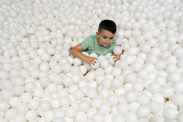 Smiling kid immersed in a pool of white plastic balls at an indoor play zone, enjoying fun and sensory playtime in a bright, modern kids attraction area.
