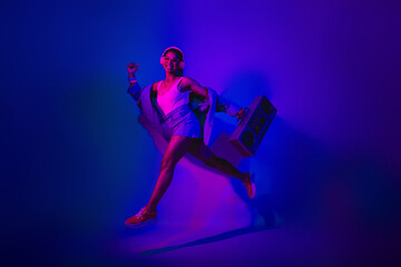 Dynamic young woman with retro boombox enjoying dance in trendy ultraviolet neon lighting for an...