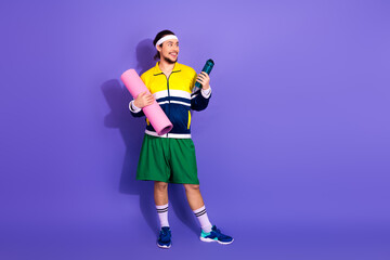 Young man in retro sportswear holding yoga mat and water bottle on purple background, embodying...