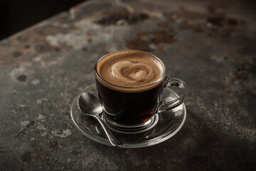 Steaming dark coffee with beautiful crema in a glass cup on a saucer with spoon. Rustic, textured surface. Warm, inviting feel for a cozy morning.