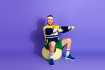 Young athletic man in retro sports outfit exercising with dumbbells on a fitness ball against a...