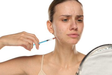 Studio shot of young woman with acne examining face in mirror and holding syringe, isolated on...