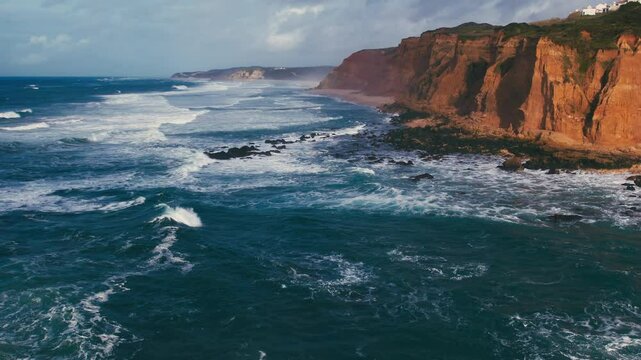 The breathtaking coastal scenery showcases the powerful ocean waves that crash with immense force against the rugged cliffs, all beneath a dramatically beautiful sky filled with clouds and sunlight
