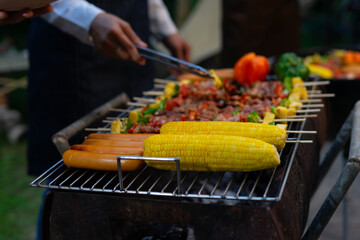 Person grilling corn sausages and skewers at outdoor barbecue