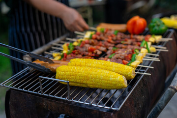 Grilled corn and skewers on barbecue grill with person cooking