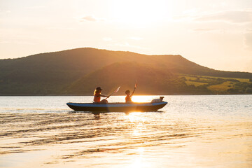 Active family lifestyle at Tsonevo Lake, Bulgaria with child paddling alongside woman and Jack Russell dog in inflatable boat across calm water and dramatic cliffs sunset summer.