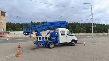 A specialized truck tower with a fenced area for safe repair work at height stands on an asphalt sidewalk near the roadway and is fenced with plastic posts. Nearby buildings, lampposts and trees