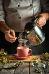 A person carefully pours steaming water from a teapot into a pink cup while straining herbs. The...