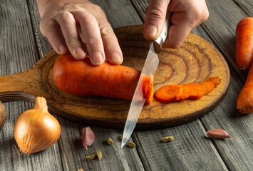 Hands skillfully cut a vibrant orange carrot on a round wooden cutting board. Nearby, are onions...
