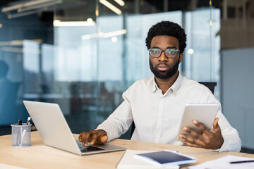 A focused African-American man working on a laptop while holding a tablet, looking directly at the...