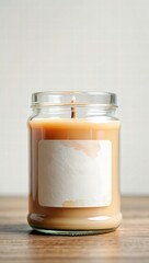 Beige wax candle in a transparent glass jar with a blank label, standing on a neutral surface with soft lighting.
