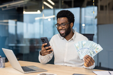 A smiling Black man in glasses holding money and looking at his phone, likely at good news in an...