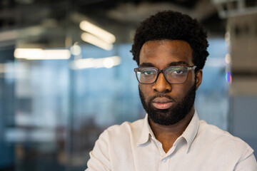 A serious man with glasses gazes intently at the camera, set against an office background.