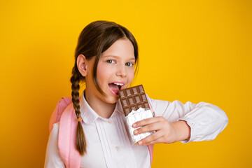 A cheerful young schoolgirl holding a chocolate bar against a yellow background, enjoying a sweet treat while dressed in uniform.