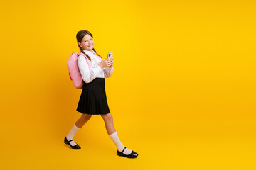 Cheerful schoolgirl wearing a uniform and holding a smartphone while walking against a yellow...