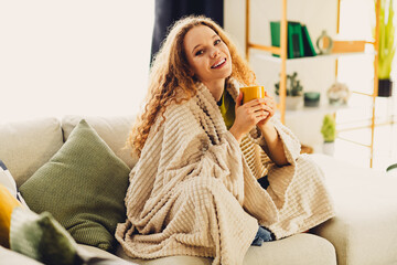Young woman with curly hair enjoying a cozy moment at home, wrapped in a blanket, holding a mug and...