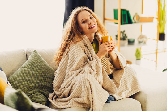 Young woman with curly hair enjoying a cozy moment at home, wrapped in a blanket, holding a mug and smiling.