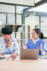 Two healthcare professionals, one male and one female, wearing medical uniforms, discuss patient care at desk with laptop, documents, and glasses in bright modern office setting