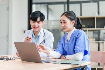 Two Asian medical professionals, male doctor and female nurse, discuss patient information while looking at laptop in modern hospital office, collaborating with focus and teamwork
