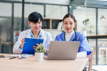 Two young Asian medical professionals, man and woman, wearing lab coats and scrubs, collaborate at desk with laptop and clipboard in modern office, showing teamwork and focus
