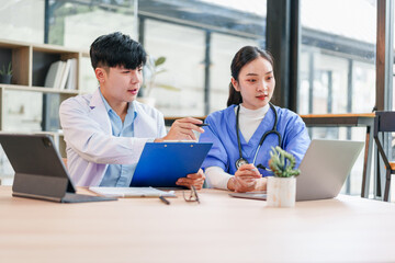 Two healthcare professionals, one male and one female, wearing medical uniforms, discuss patient information with clipboard and laptop in modern office, showing teamwork and focus