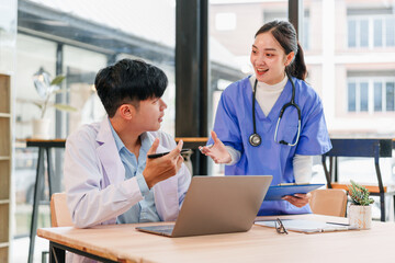Two healthcare professionals, one male and one female, discuss patient information at desk with laptop, clipboard, and coffee cup in bright, modern medical office, expressing teamwork and focus