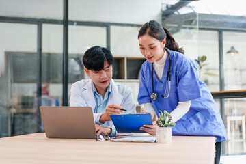 Two healthcare professionals, Asian male doctor and Asian female nurse, discuss patient information with focus and teamwork in modern hospital office, using laptop and clipboard