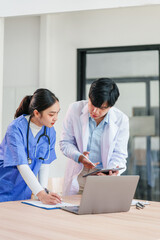 Two healthcare professionals, one in blue scrubs and one in white coat, collaborate at desk with laptop, clipboard, and tablet, showing teamwork and focus in modern medical office