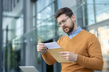 A worried young man sits on a bench near an office building and reads a letter he received in the...