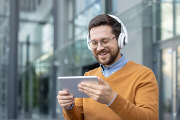 Close-up photo of a young man wearing headphones sitting on a bench outside and using a tablet