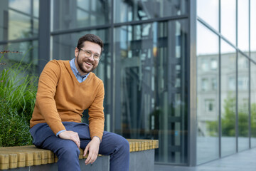 Portrait of a smiling young businessman sitting on a bench near an office building and looking at the camera