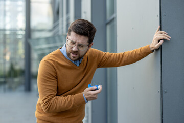 Leaning against the wall of a building, a young man suffers from pain and shortness of breath, using an asthma inhaler
