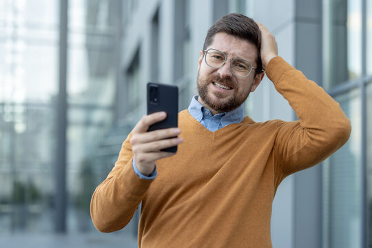 Close-up photo of a young stressed businessman standing outside his office building, holding his phone, holding his head in his hand and looking at the camera sadly
