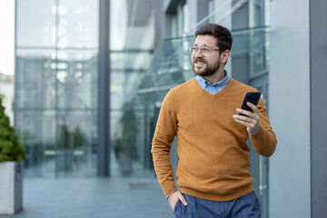 A young businessman is walking near an office building outside, holding a mobile phone in his hand and looking to the side