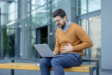 A young businessman, twisted in pain, sits on a bench outside near a building, working on a laptop,...