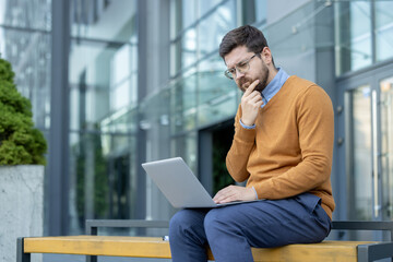A thoughtful young man sits on a bench on the street near an office building and looks intently at the laptop screen