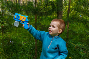 A smiling child holds a bubble gun in his hands while playing in nature. Soap bubbles.