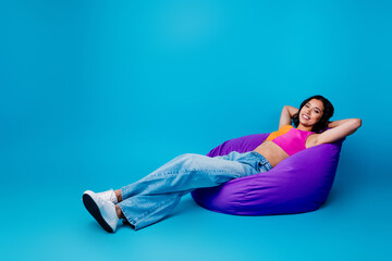Young woman relaxing on a purple bean bag against a blue background in casual and stylish summer...