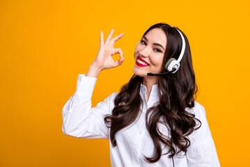 Young cheerful female call center worker wearing headset making OK hand sign against yellow background