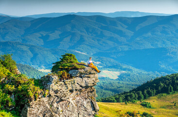 Man relaxing in sleeping bag on mountain cliff, enjoying scenic sunrise view over forested hills