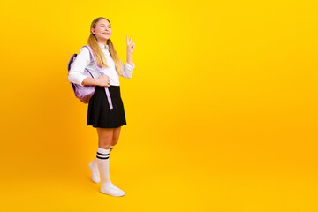 Cheerful schoolgirl with a backpack showing peace sign on vibrant yellow background